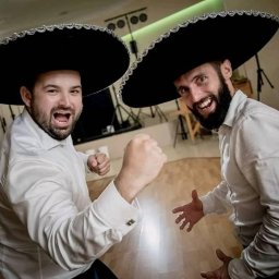 Waldemar Kozielewski - Dwóch mężczyzn w białych koszulach i czarnych sombrerach, jeden z zaciśniętą pięścią, obaj uśmiechnięci, na tle sprzętu oświetleniowego.