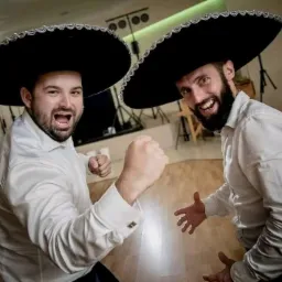 Dwóch mężczyzn w białych koszulach i czarnych sombrerach, jeden z zaciśniętą pięścią, obaj uśmiechnięci, na tle sprzętu oświetleniowego.