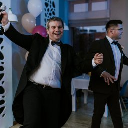 Waldemar Kozielewski - Rozbawiony wodzirej z mikrofonem i otwartą marynarką, prowadzący zabawę podczas wesela, w tle drugi mężczyzna w garniturze i dekoracje z balonów.