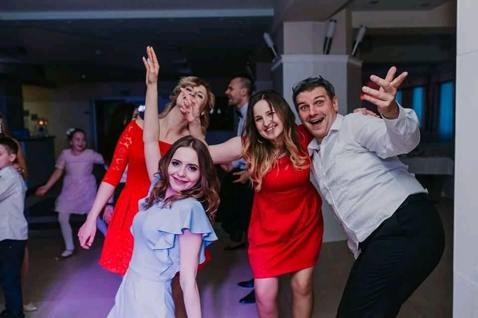 Rozbawieni goście na parkiecie podczas imprezy, kobiety w czerwonych i niebieskich sukienkach, mężczyzna w białej koszuli i czarnych spodniach, dynamiczne pozy.