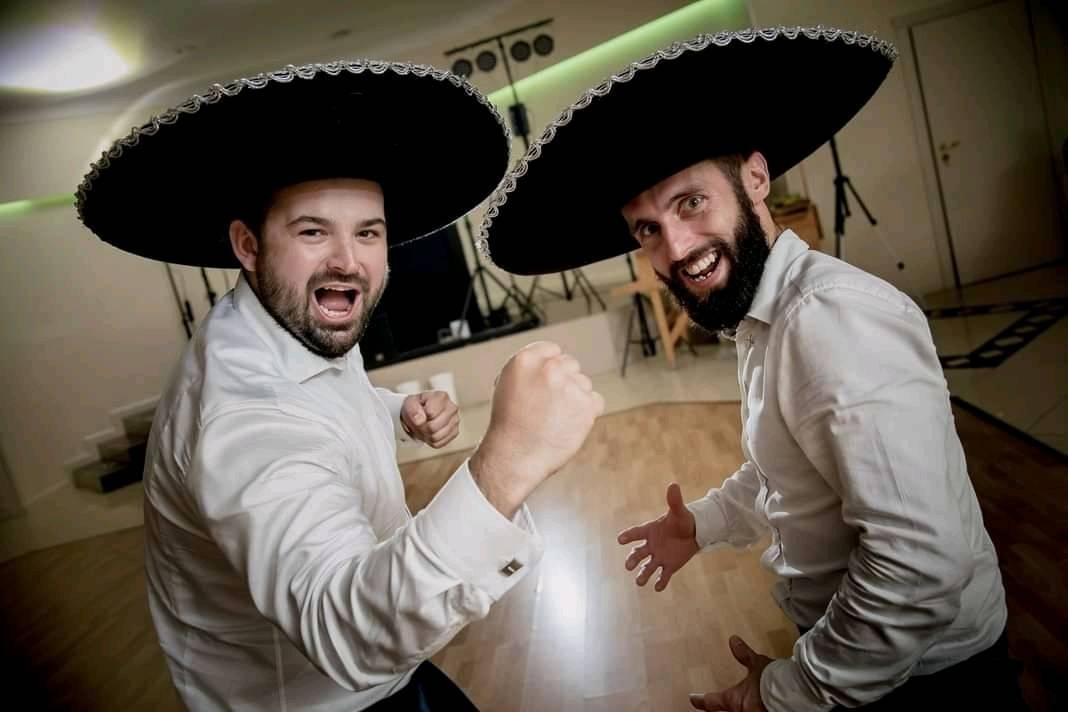 Dwóch mężczyzn w białych koszulach i czarnych sombrerach, jeden z zaciśniętą pięścią, obaj uśmiechnięci, na tle sprzętu oświetleniowego.