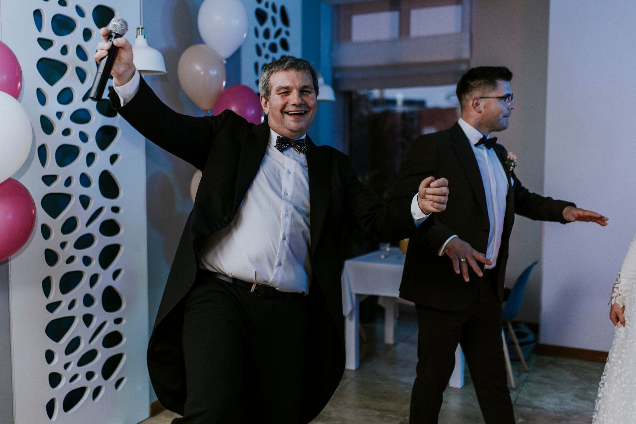 Rozbawiony wodzirej z mikrofonem i otwartą marynarką, prowadzący zabawę podczas wesela, w tle drugi mężczyzna w garniturze i dekoracje z balonów.