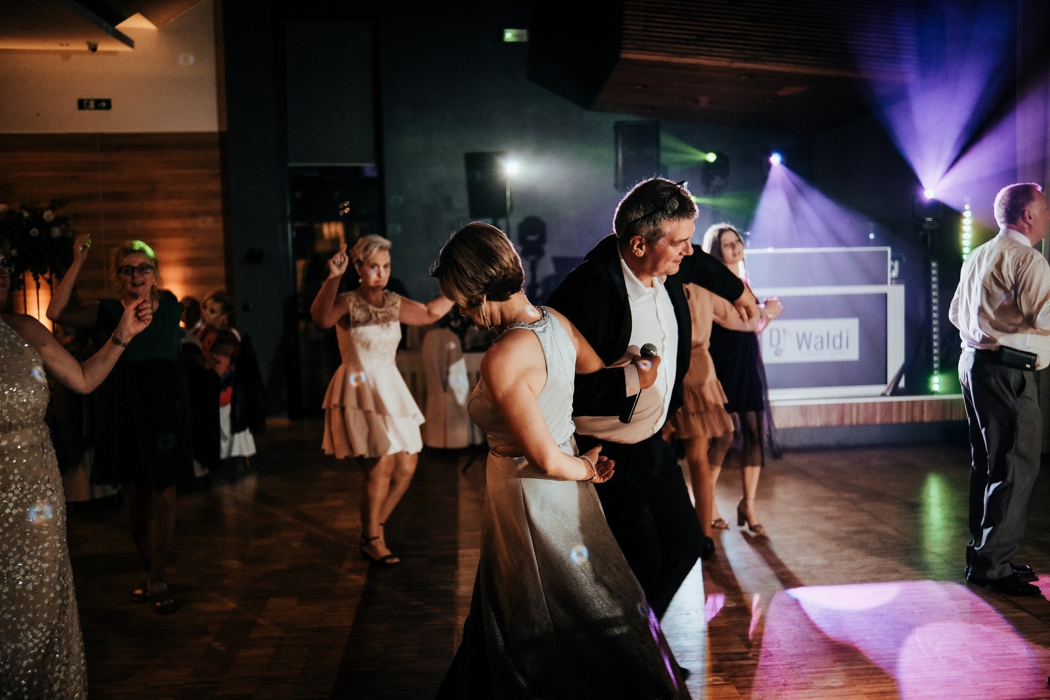 Parkiet taneczny podczas uroczystości, w tle oświetlenie sceniczne i DJ z mikrofonem w dłoni, tańczący z gośćmi.
