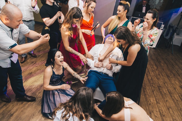 Rozbawiony mężczyzna z zawiązanymi oczami, otoczony przez roześmiane kobiety i mężczyzn podczas imprezy, parkiet z desek w tle.