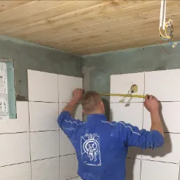 Pracownik w niebieskiej bluzie z nadrukiem mierzy odległość między płytkami na ścianie w łazience, widoczne krzyżyki dystansowe, surowy sufit z desek i okno.