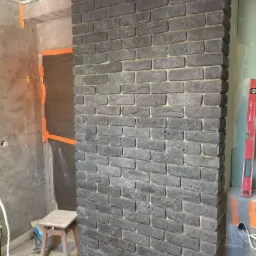 Fragment ściany wykończonej szarą cegłą dekoracyjną w trakcie prac wykończeniowych, widoczna poziomica i fragment surowego wnętrza z profilem sufitowym.