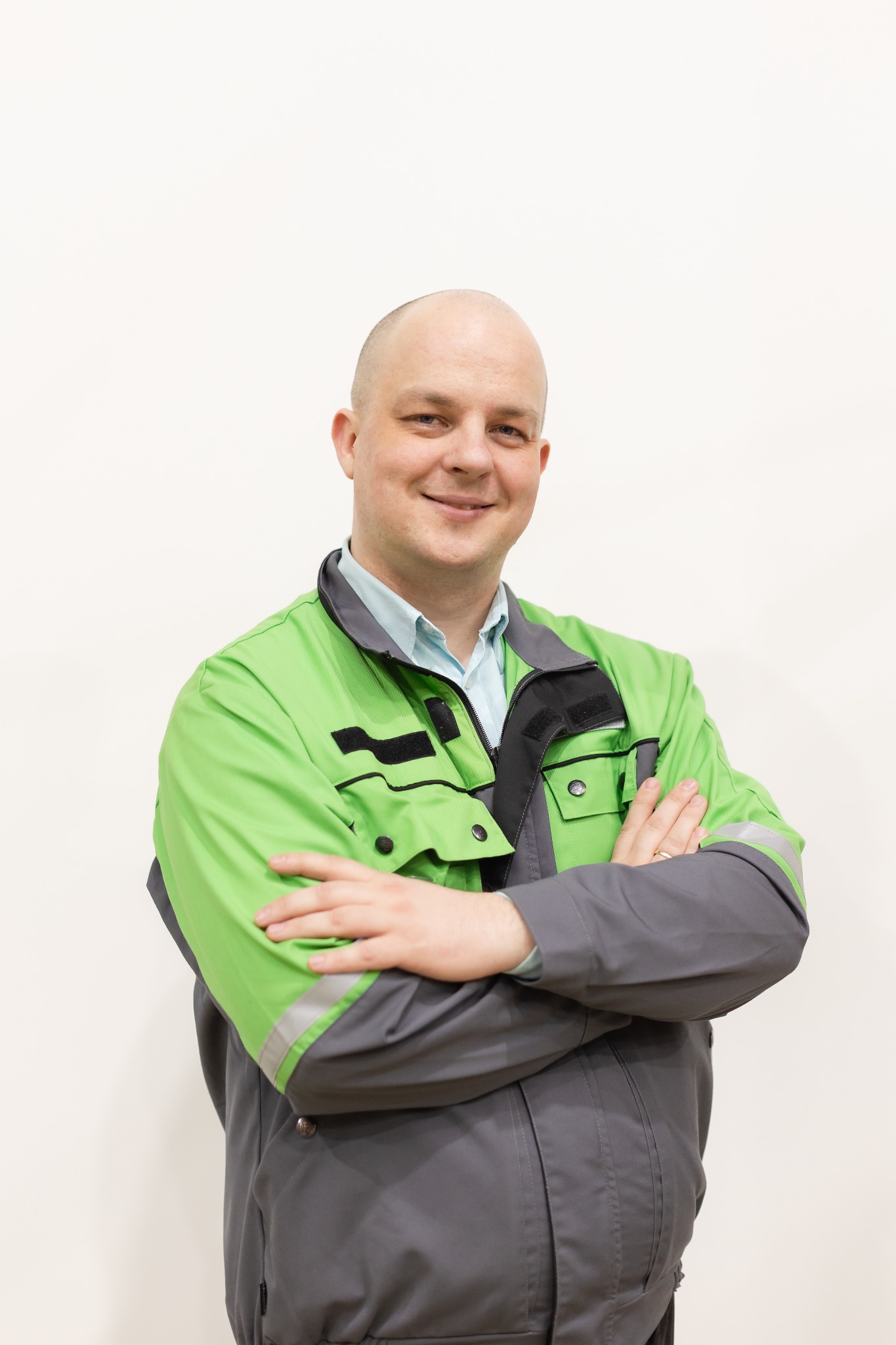 Portret uśmiechniętego mężczyzny w zielono-szarym stroju roboczym, z założonymi rękami na tle jasnej ściany. Widoczny pierścionek na palcu.