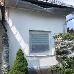 Przygotowanie do odświeżenia elewacji: oklejone folią okno, rynna i krzewy, świeżo pomalowana ściana domu.