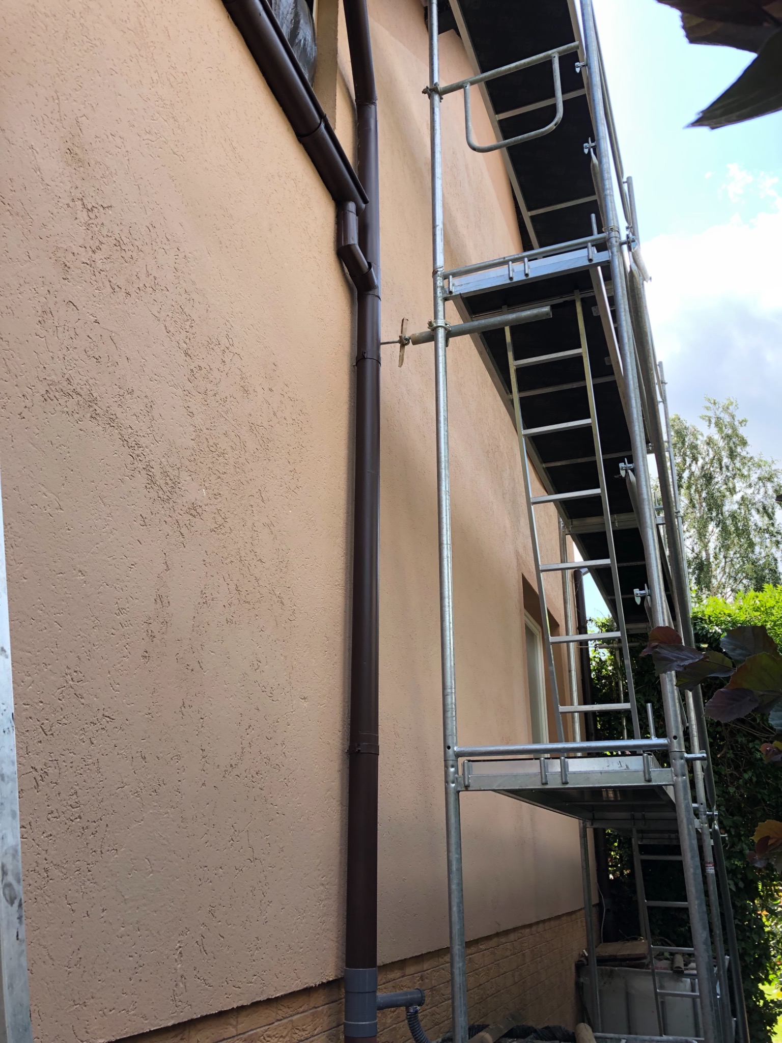 Fragment elewacji budynku z beżowym tynkiem strukturalnym, obok brązowa rynna i metalowe rusztowanie.