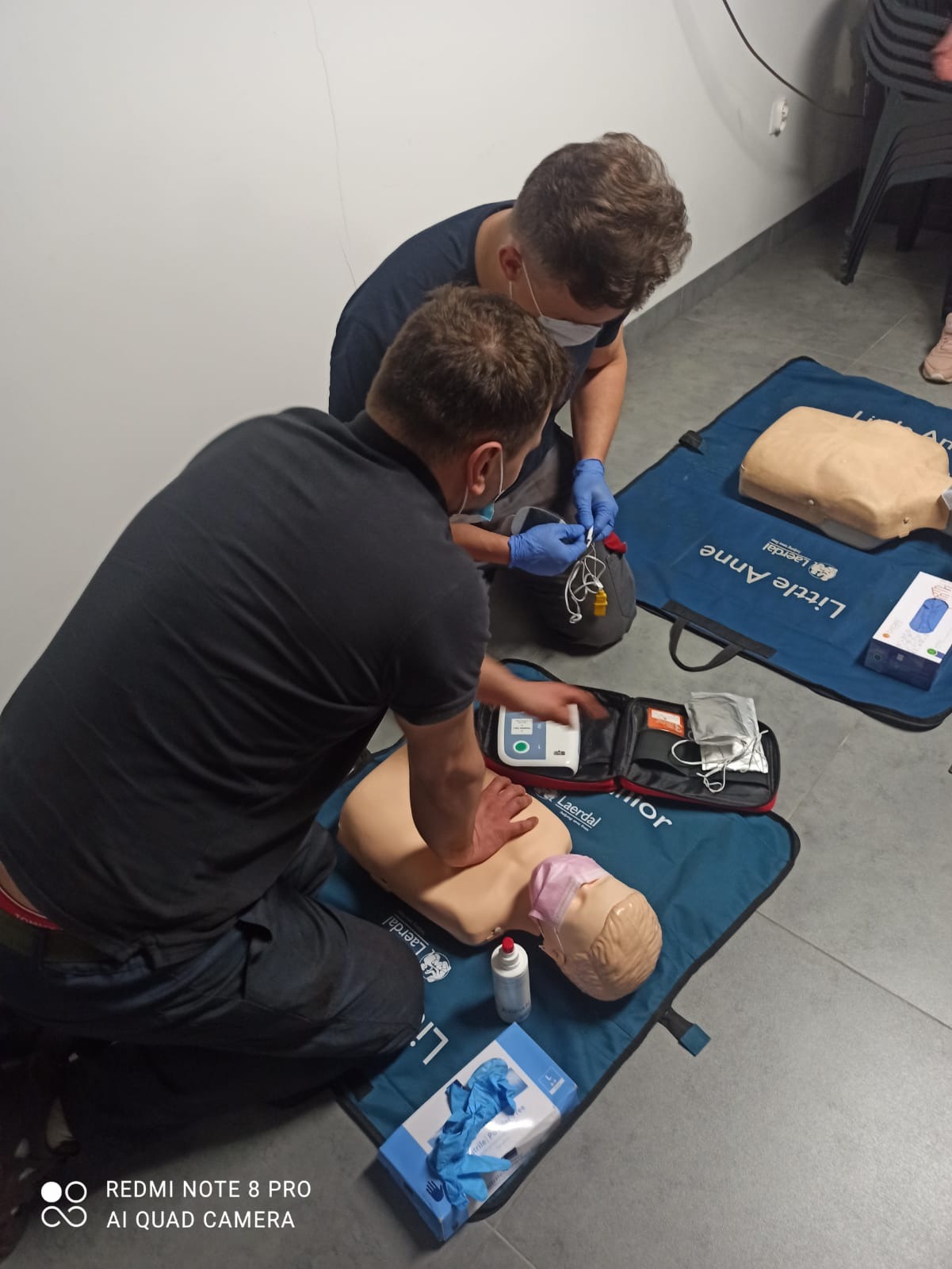 Instruktor demonstruje resuscytację krążeniowo-oddechową na manekinie Little Anne, drugi instruktor przygotowuje defibrylator AED, obaj w maseczkach i rękawiczkach, na podłodze rozłożone maty Laerdal.