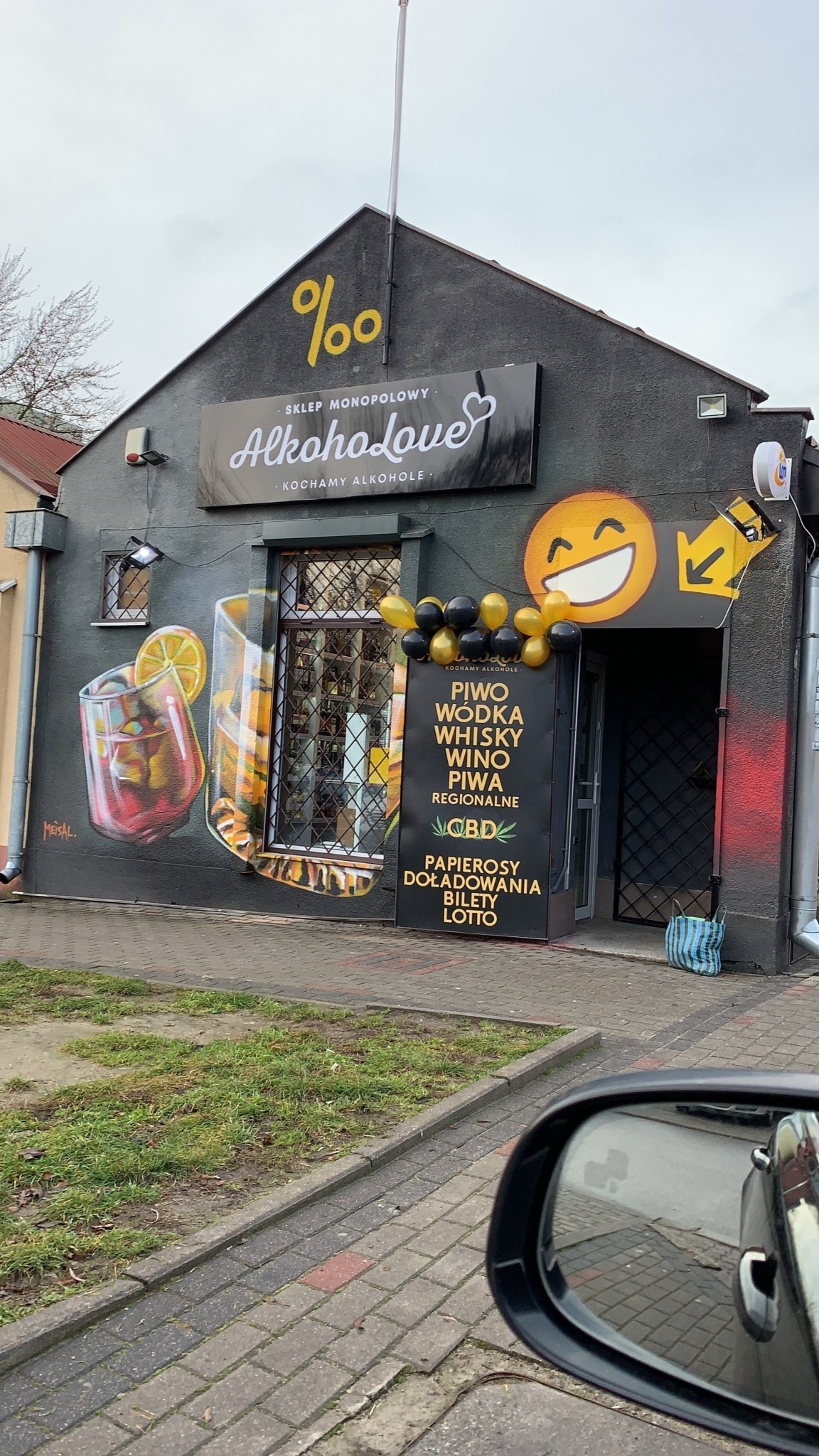 Fasada sklepu monopolowego z muralami przedstawiającymi drinki, szyldem 'AlkohoLove' i tablicą informującą o dostępnych produktach, udekorowana czarno-złotymi balonami.