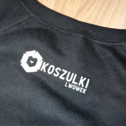 Fragment czarnej koszulki z białym nadrukiem 'KOSZULKI LWOWEK' i graficznym logo w kształcie sześcioboku z elementami trójkątnymi w środku, leżącej na drewnianej powierzchni.