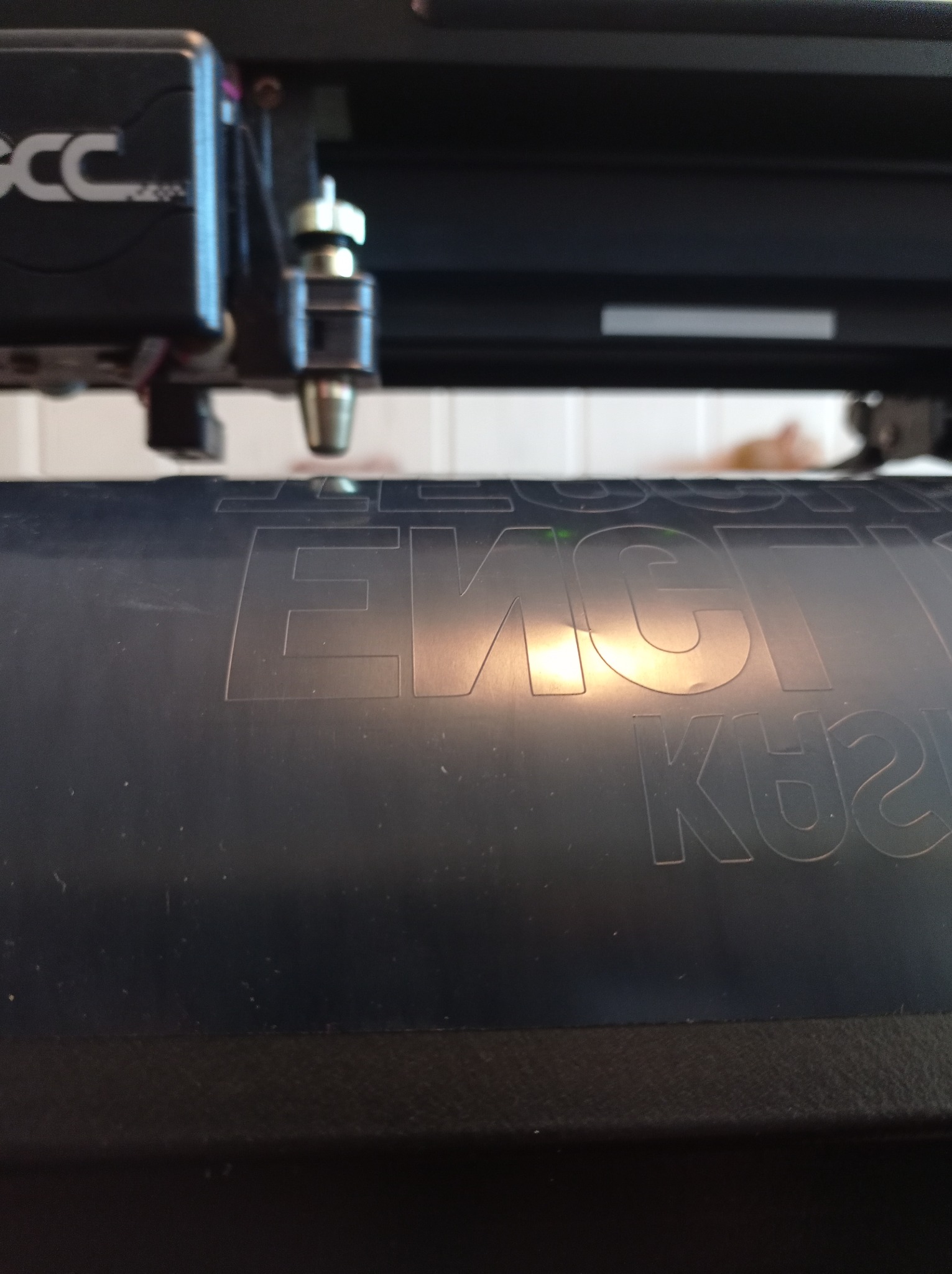 Zbliżenie na ploter tnący GCC podczas pracy, widoczne wycięte litery w czarnym materiale, delikatne odbicie światła na powierzchni.