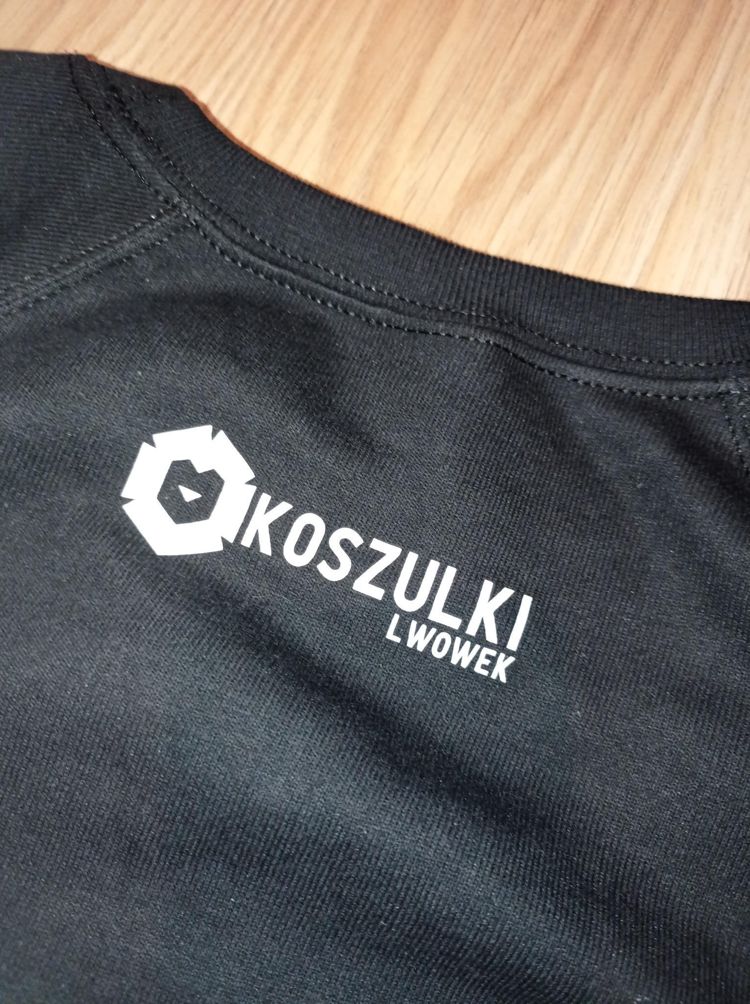 Fragment czarnej koszulki z białym nadrukiem 'KOSZULKI LWOWEK' i graficznym logo w kształcie sześcioboku z elementami trójkątnymi w środku, leżącej na drewnianej powierzchni.