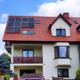 Dwupiętrowy dom z czerwonym dachem i panelami słonecznymi na dachu, widok z dołu, błękitne niebo z chmurami w tle.