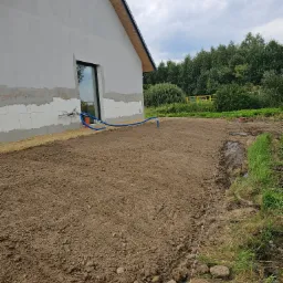 Przygotowany teren pod trawnik przy nowym domu. Widoczne nawodnienie i wyrównana ziemia. W tle budynek w trakcie wykończenia i drzewa.