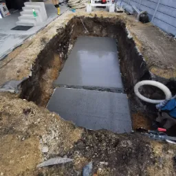 Wykopany dół z wylanym betonem na dnie, widoczne ściany wykopu z ziemi, w tle elementy ogrodu i ogrodzenia.