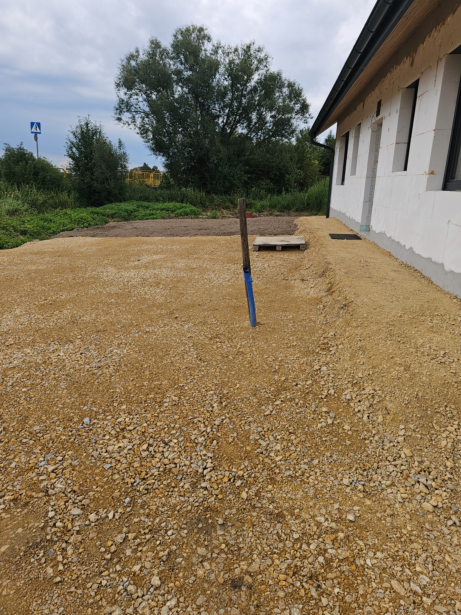 Teren po niwelacji, wyrównany żwirem przy budynku z pustaków. W tle zieleń i drzewa. Widoczny słupek z niebieską rurą i paleta.