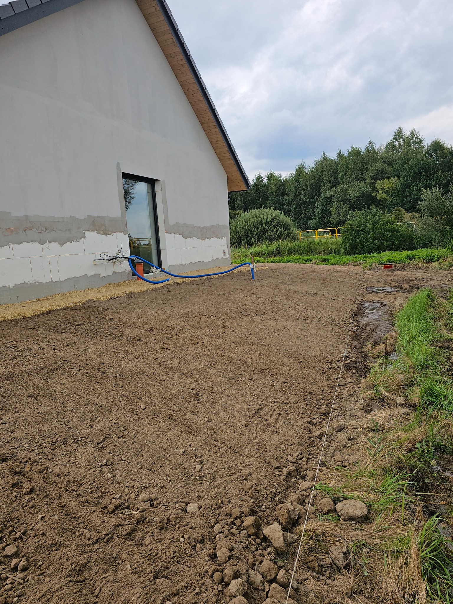 Przygotowany teren pod trawnik przy nowym domu. Widoczne nawodnienie i wyrównana ziemia. W tle budynek w trakcie wykończenia i drzewa.
