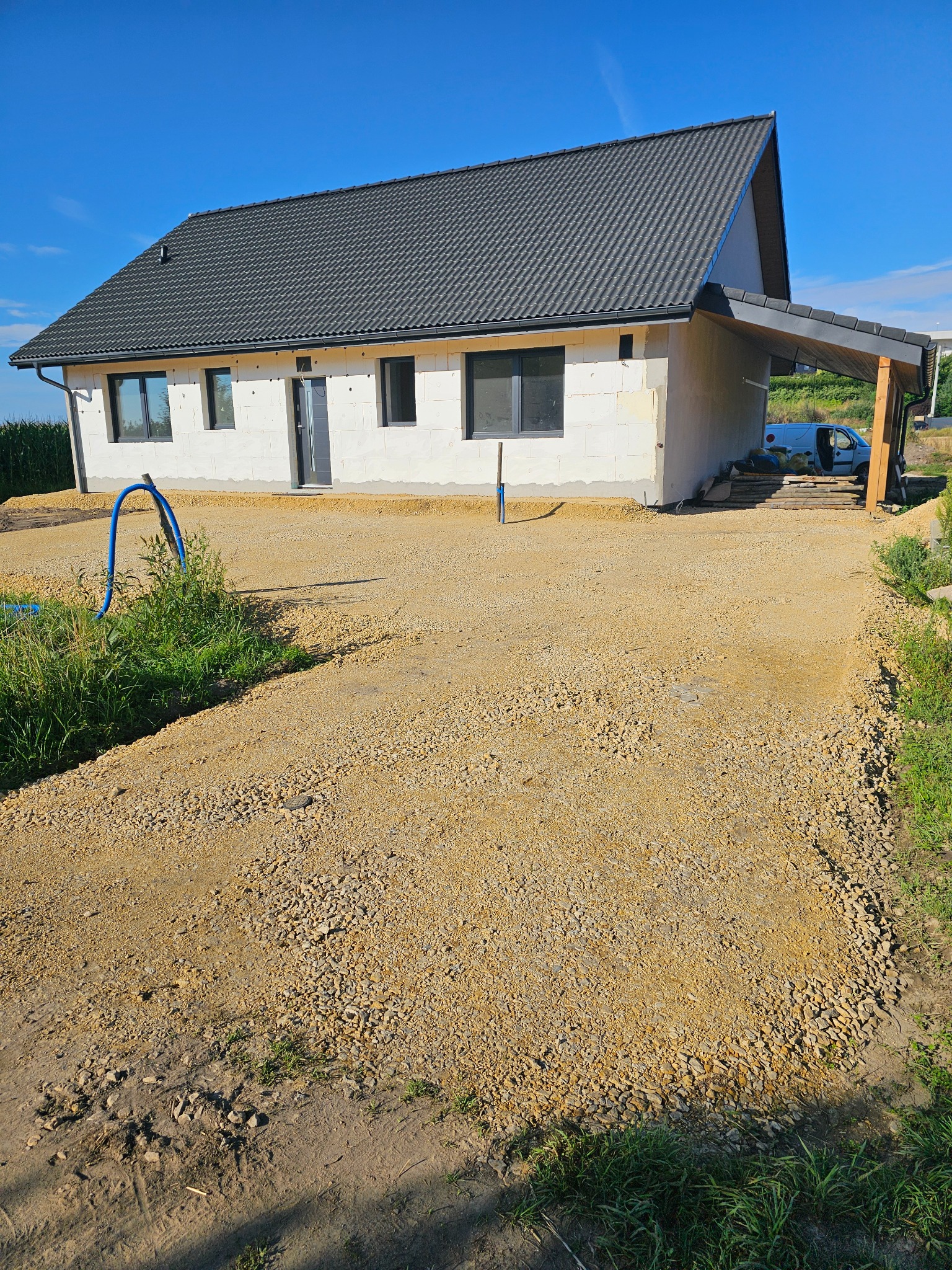 Dom w trakcie budowy z utwardzonym terenem wokół. Widoczny gruz i piasek. W tle błękitne niebo i zieleń. Dom z szarym dachem i białymi ścianami z pustaków.