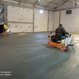 Profest Posadzki Przemysłowe - Dwaj operatorzy zacierają świeżo wylaną posadzkę w hali przemysłowej za pomocą zacieraczek mechanicznych, widoczne ślady po pracy maszyn.