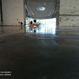 Profest Posadzki Przemysłowe - Błyszcząca, szara podłoga w hali produkcyjnej, z widocznymi śladami polerowania i maszyną do szlifowania w tle, przy otwartych wrotach hali.