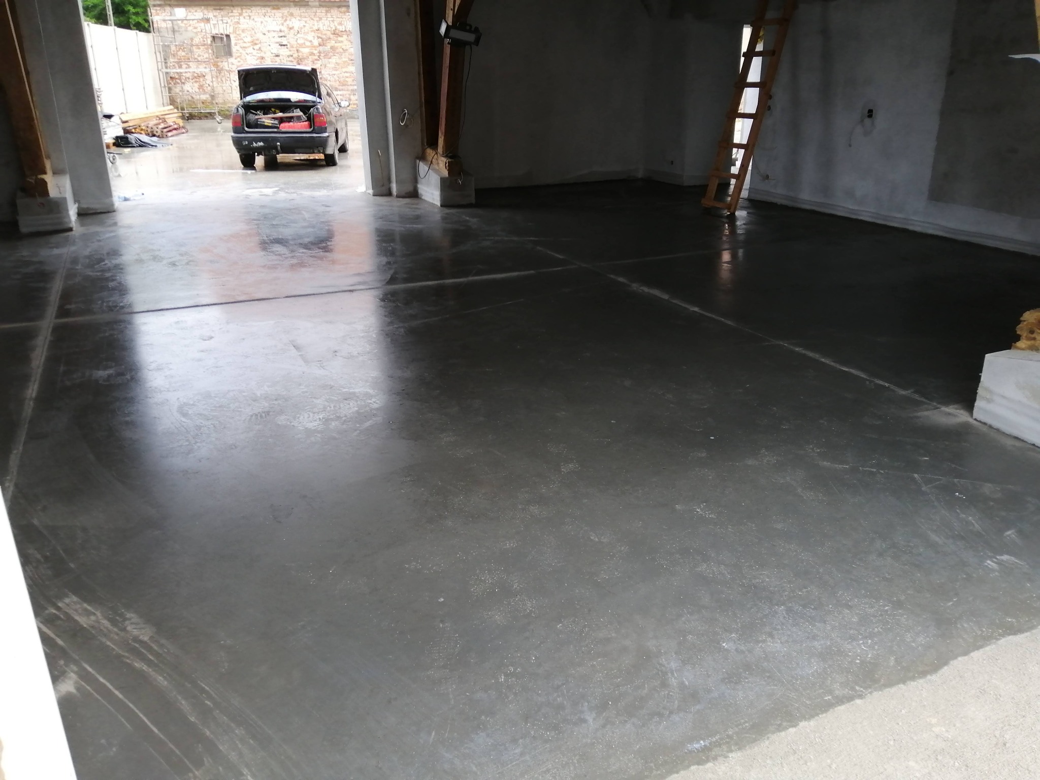 Świeżo wylana, gładka, betonowa podłoga wewnątrz budynku, widoczna drabina oparta o ścianę i samochód z otwartym bagażnikiem na zewnątrz budynku.