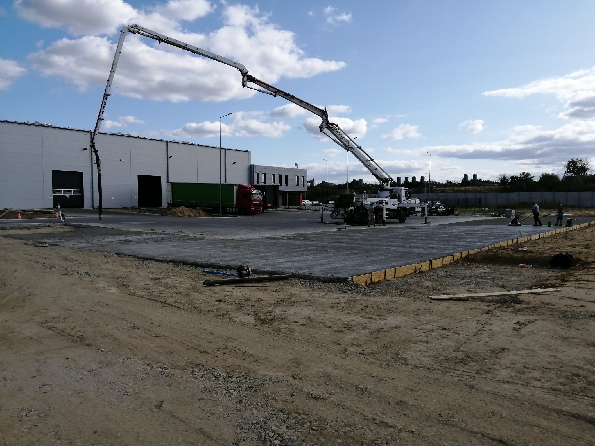 Wylewanie betonu na plac budowy z użyciem pompy do betonu ABET, w tle hala przemysłowa i ciężarówka.