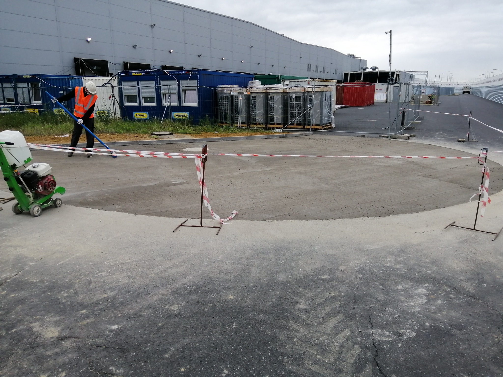 Pracownik w kamizelce odblaskowej i kasku wygładza świeżo wylaną, okrągłą powierzchnię betonową na zewnątrz budynku przemysłowego, obok stoi zielona maszyna do wygładzania betonu, teren oddzielony...