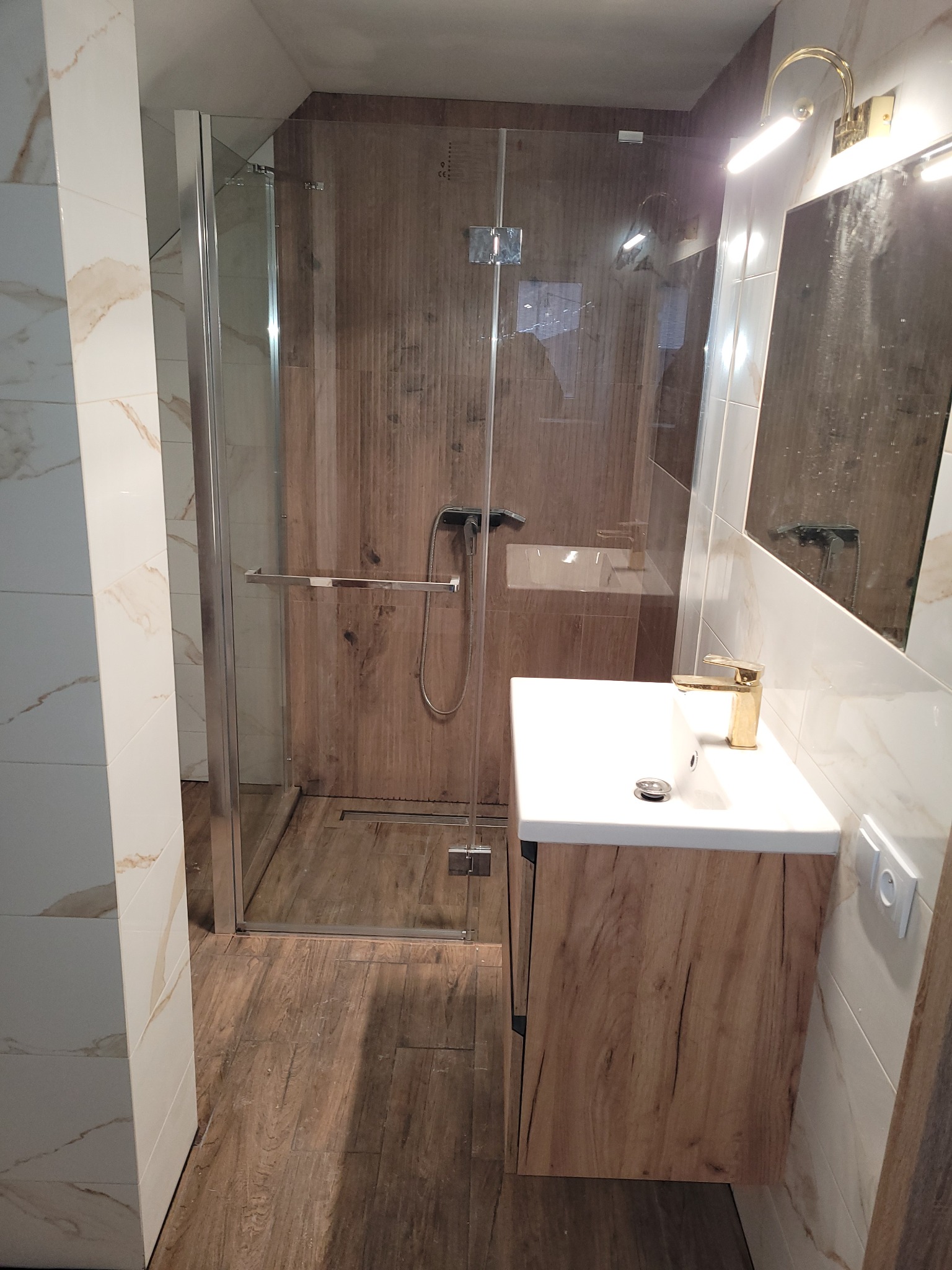 Nowoczesna łazienka z kabiną prysznicową typu walk-in, drewnopodobnymi płytkami na ścianach i podłodze, białą umywalką z szafką w kolorze drewna oraz złotą baterią.