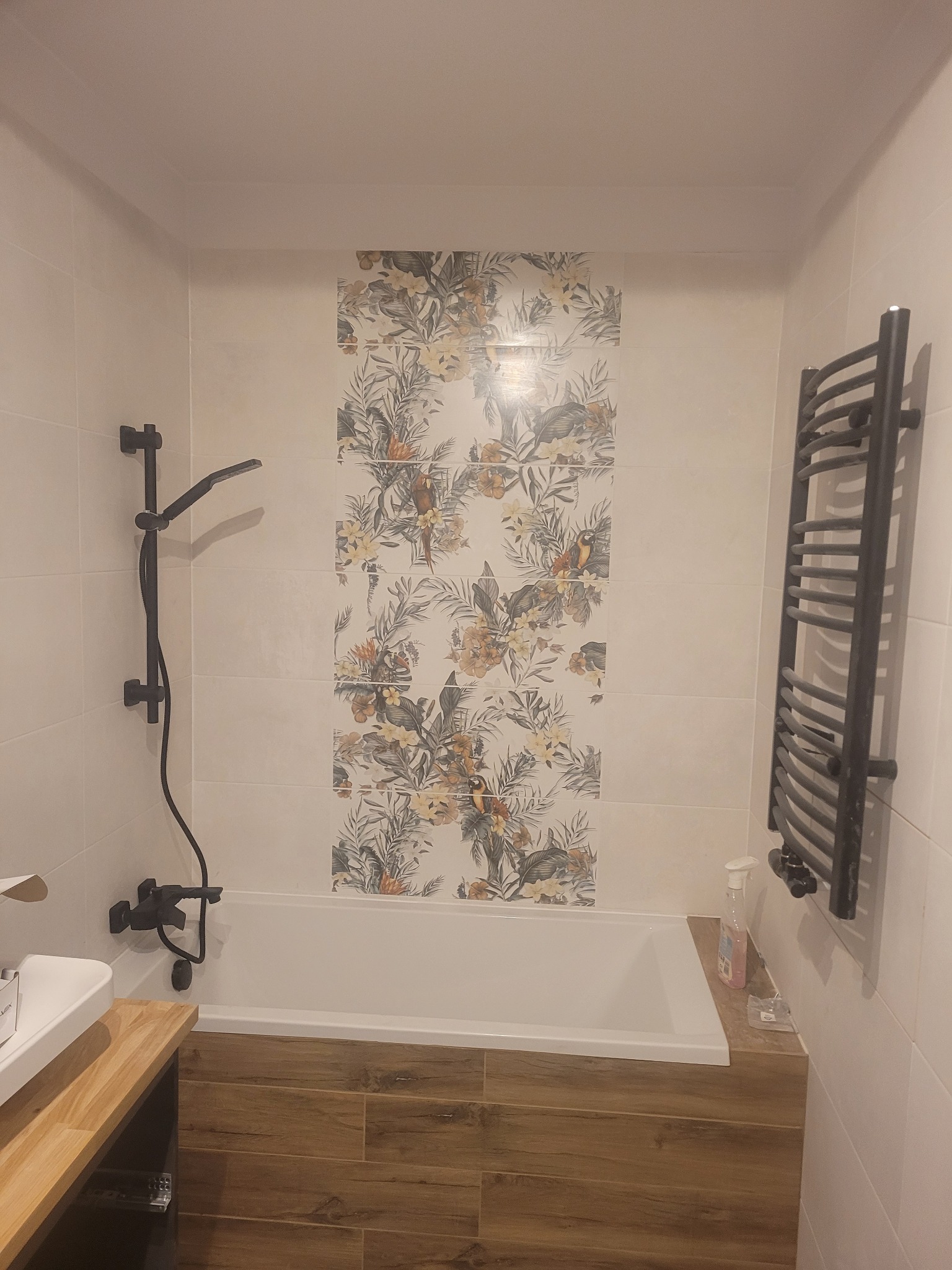 Nowoczesna łazienka z białymi płytkami, dekoracyjnym panelem z motywem tropikalnym, czarną armaturą i grzejnikiem drabinkowym, oraz zabudową wanny imitującą drewno.