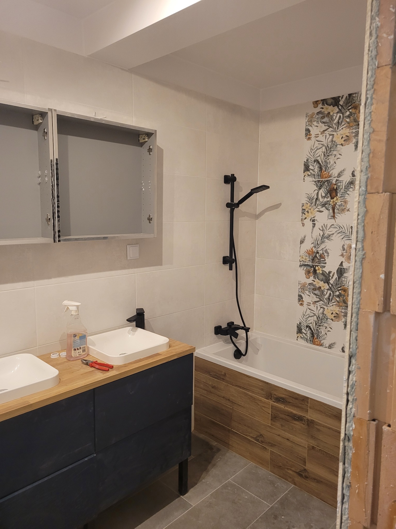 Nowoczesna łazienka z dwoma białymi umywalkami na drewnianym blacie i szafką w kolorze granatowym, czarna armatura, wanna obudowana płytkami imitującymi drewno oraz fragment ściany z dekoracyjnymi...