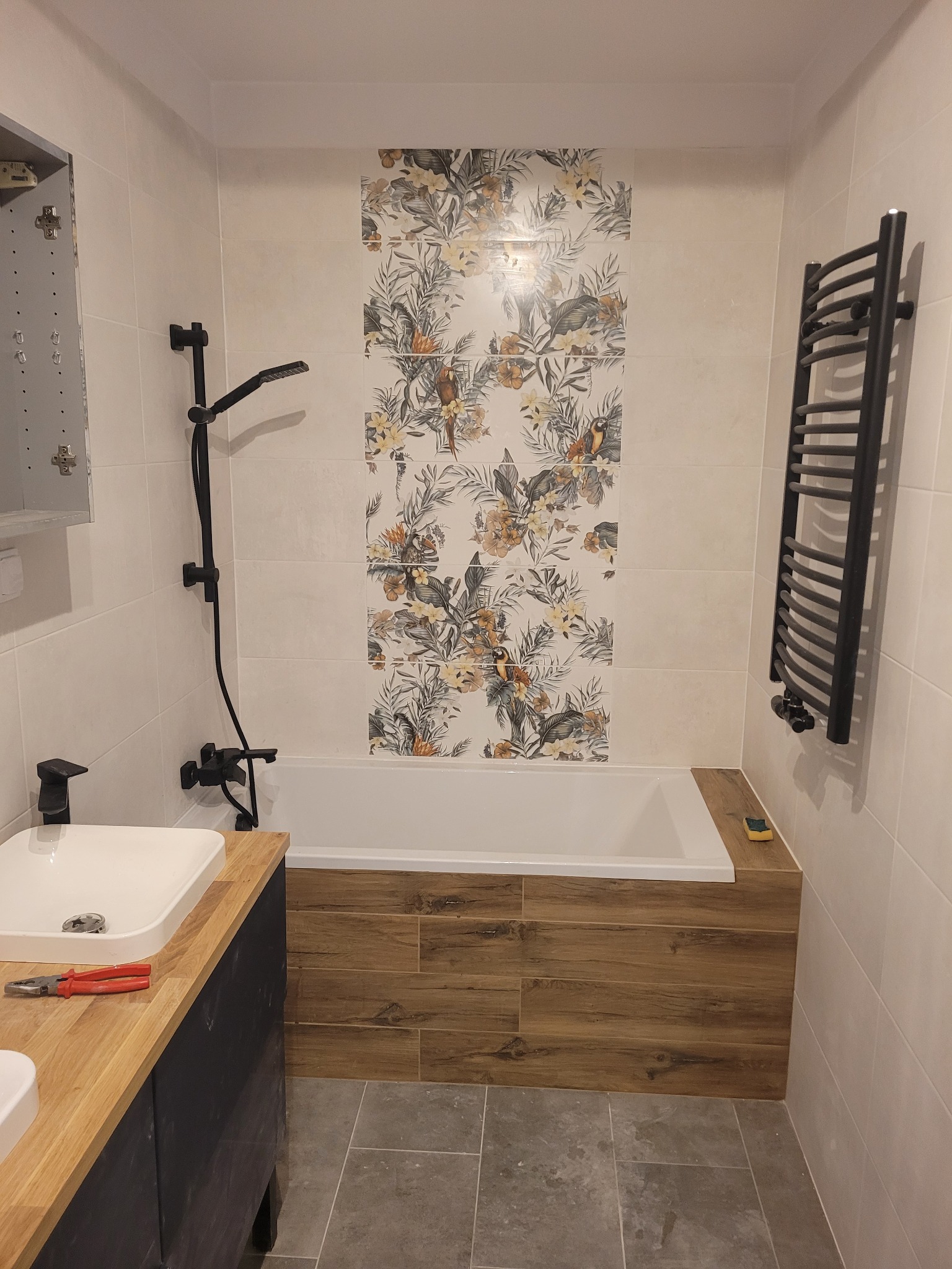 Nowoczesna łazienka z wanną obudowaną drewnopodobnymi panelami, ozdobną ścianą z motywem roślinnym, czarną armaturą i grzejnikiem drabinkowym, częściowo zamontowana szafka z lustrem oraz szczypce...