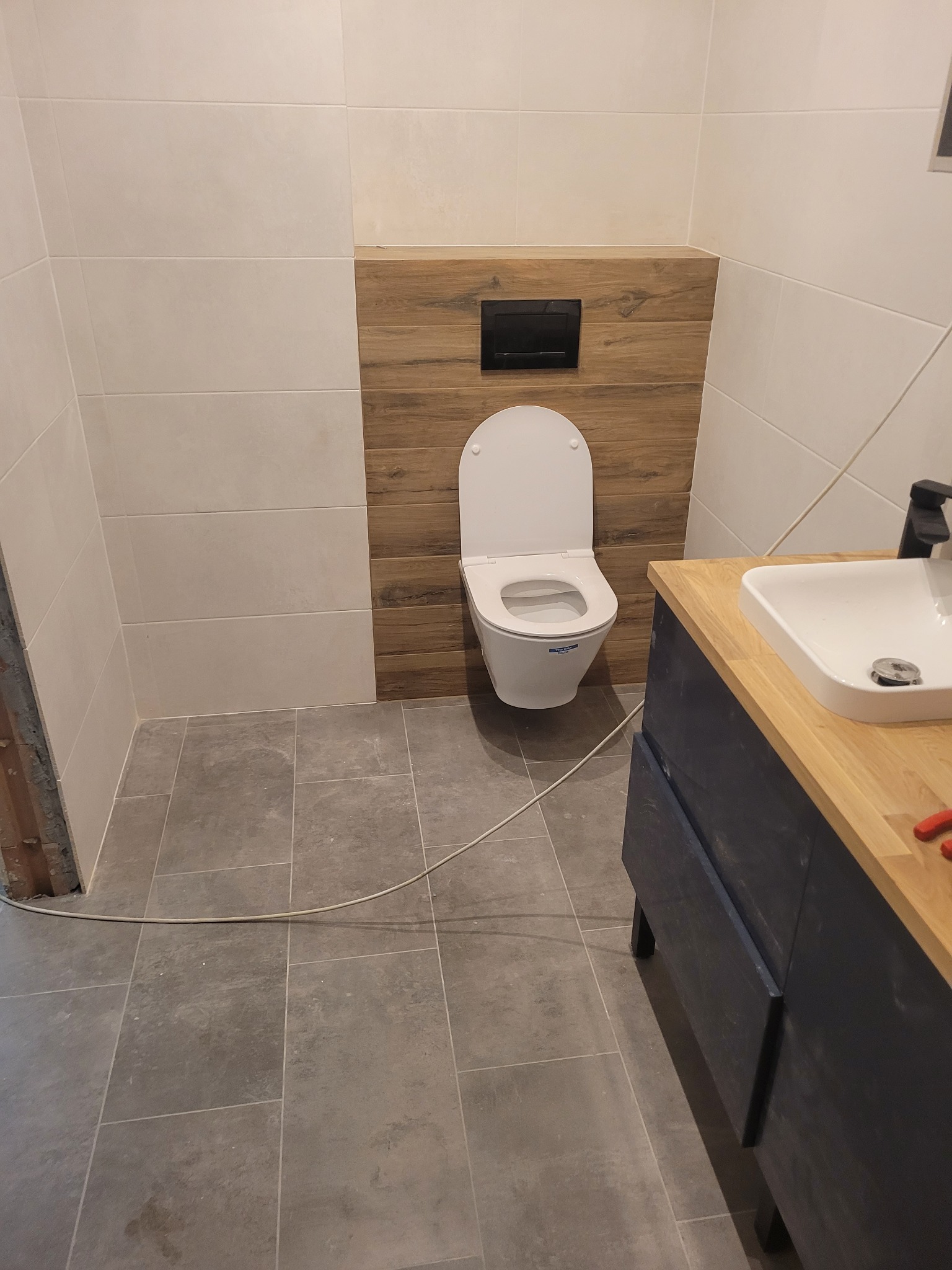 Nowoczesna łazienka z podwieszaną toaletą na tle drewnopodobnych płytek, szare płytki podłogowe i umywalka nablatowa z szafką w kolorze granatowym, widoczne przewody elektryczne w rogu.