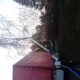 Wiercenie studni głębinowej: strumień wody pod ciśnieniem wypływa z rury wiertniczej, widoczny fragment czerwonej plandeki ciężarówki i drzewa w tle.