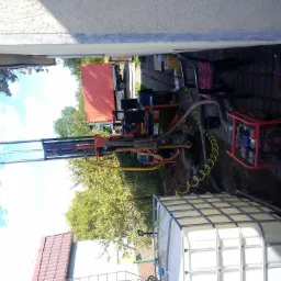 Wiercenie studni przy domu: wiertnica na podwórku, zbiornik na wodę, przewody i narzędzia. Widoczny samochód ciężarowy w tle.