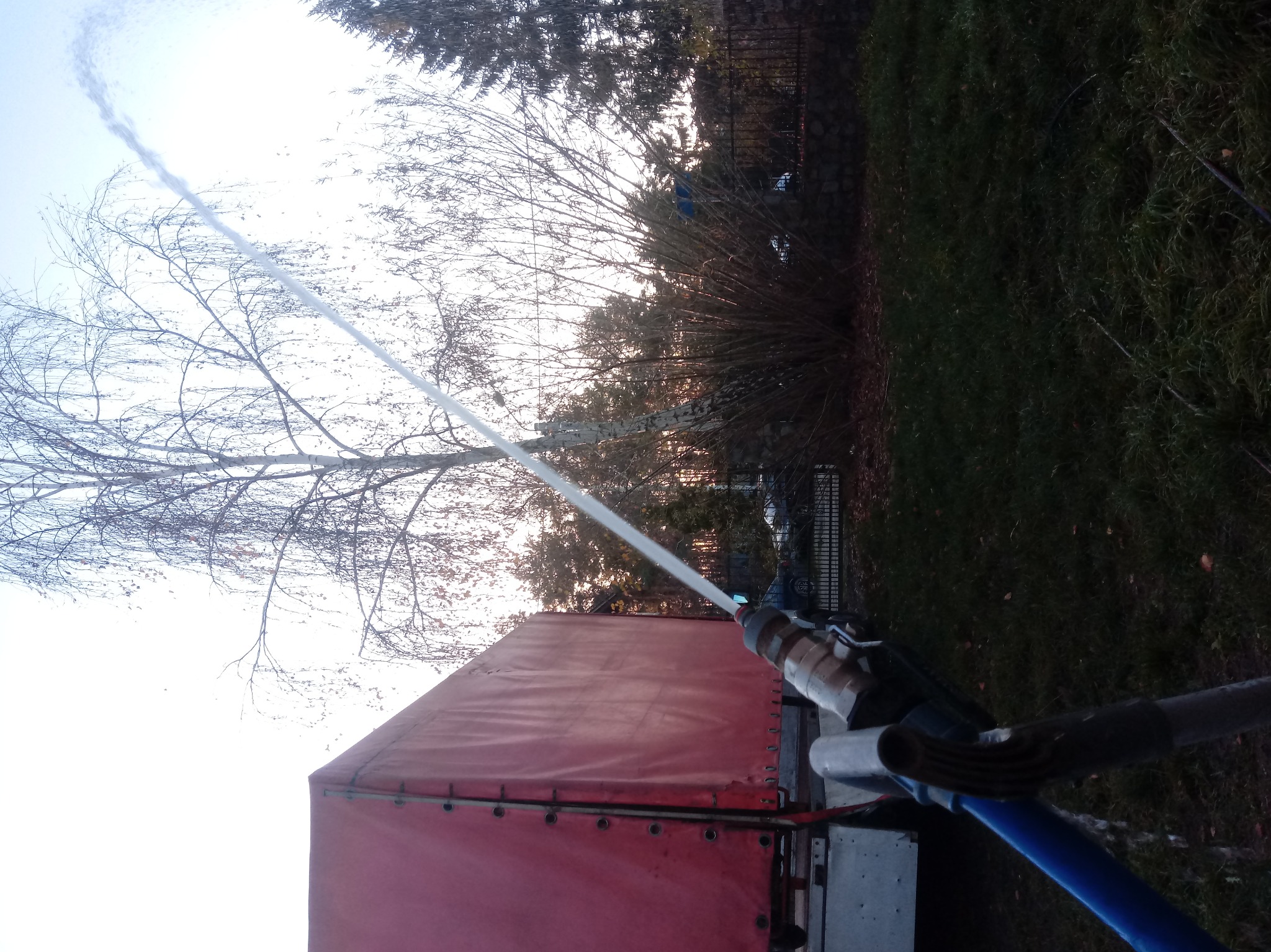 Wiercenie studni głębinowej: strumień wody pod ciśnieniem wypływa z rury wiertniczej, widoczny fragment czerwonej plandeki ciężarówki i drzewa w tle.