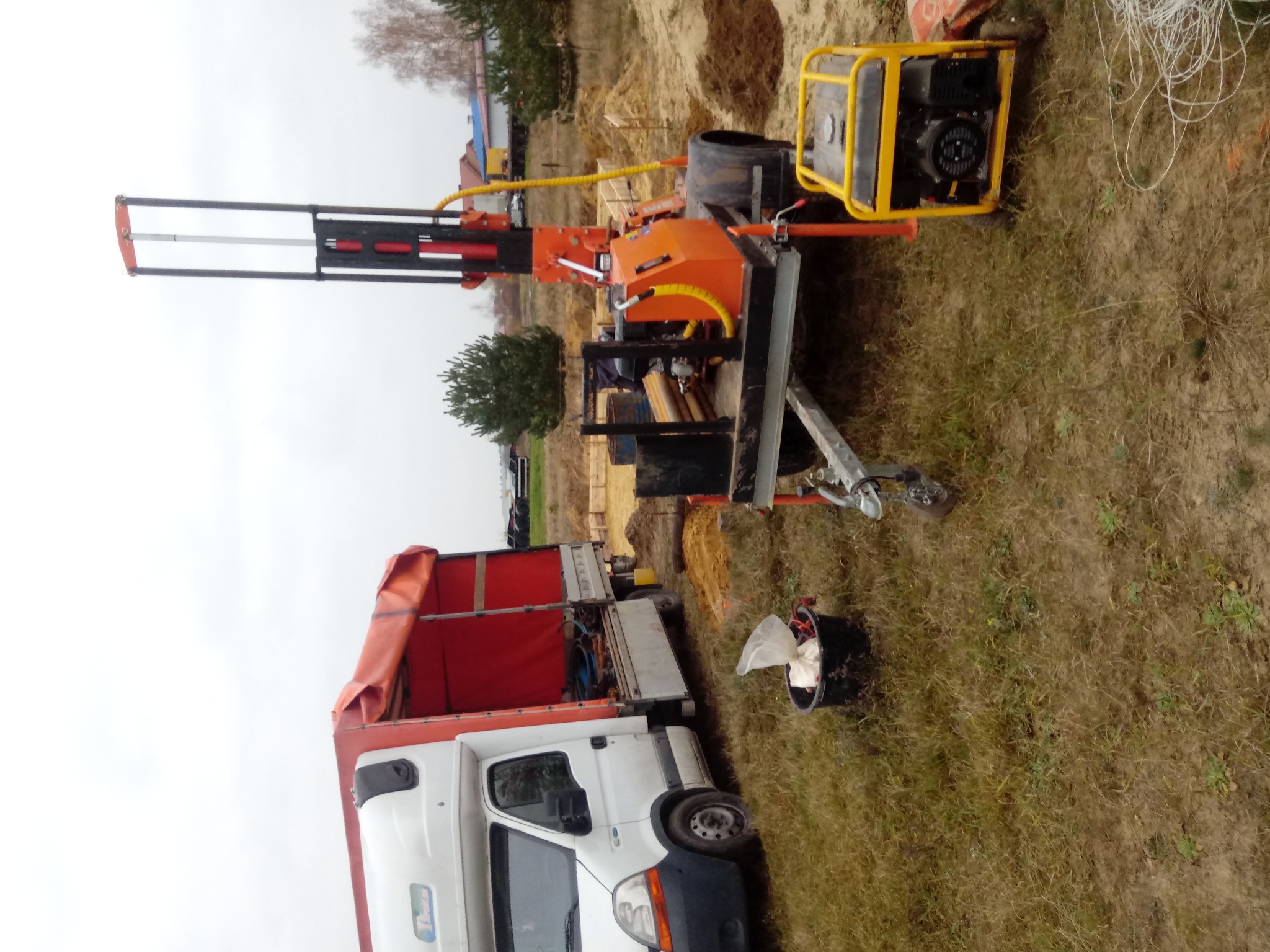 Mobilna wiertnica do studni na przyczepie za samochodem dostawczym, na trawiastym terenie z widocznym agregatem prądotwórczym i wykopem w tle.