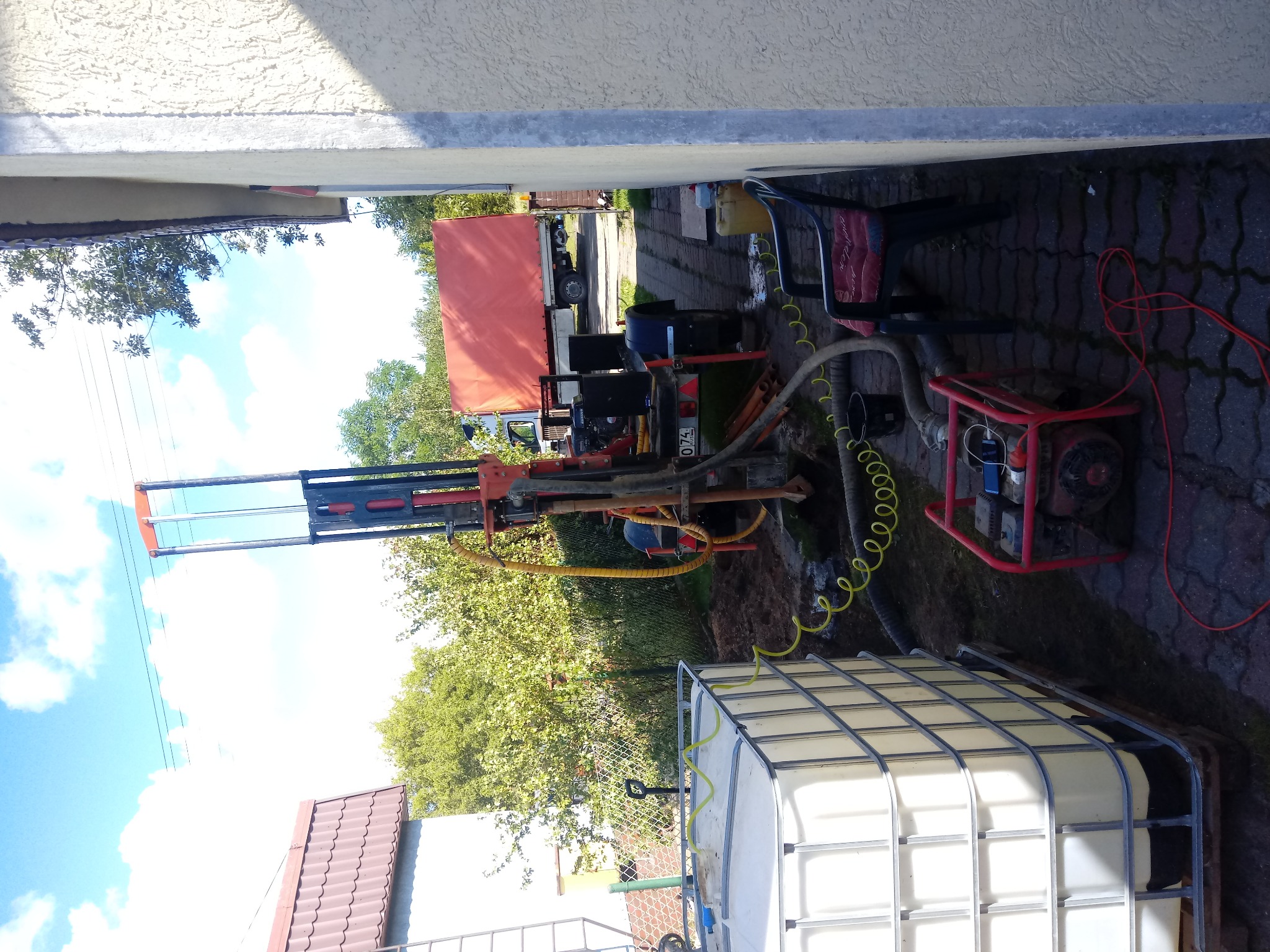 Wiercenie studni przy domu: wiertnica na podwórku, zbiornik na wodę, przewody i narzędzia. Widoczny samochód ciężarowy w tle.