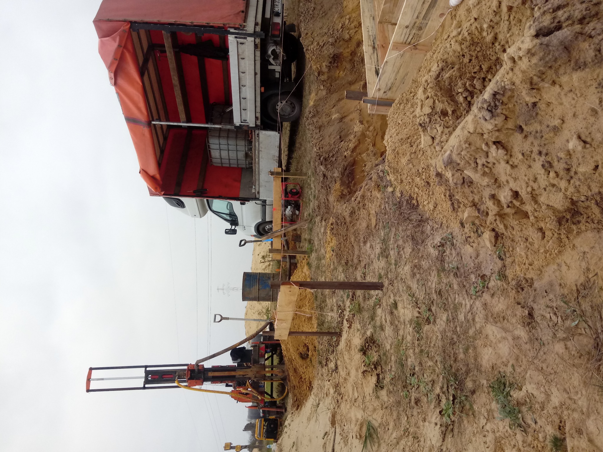 Wiercenie studni w gruncie piaszczystym, widoczna wiertnica, wykop zabezpieczony drewnianą konstrukcją, obok zaparkowany samochód ciężarowy.