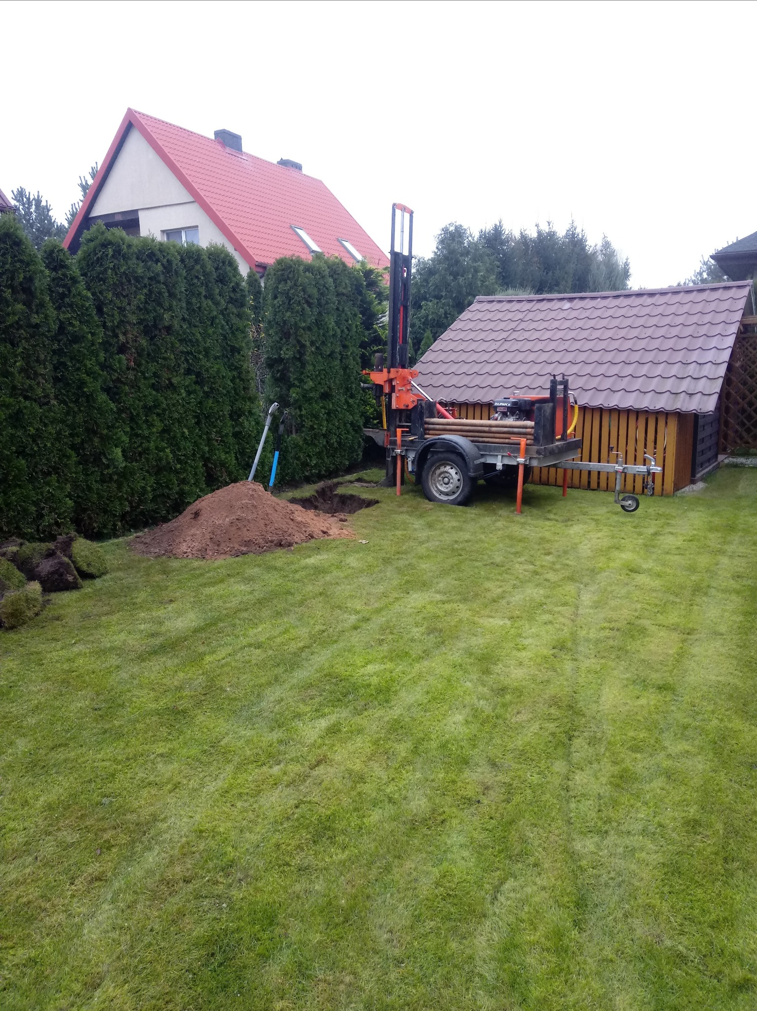 Wiercenie studni na trawniku: mobilna wiertnica obok wykopu z hałdą ziemi, w tle dom jednorodzinny z czerwoną dachówką i zielone tuje.