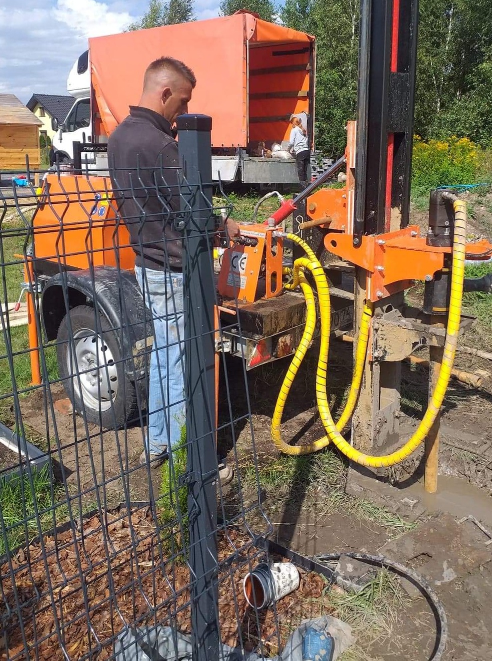 Pracownik obsługuje pomarańczową wiertnicę do studni na przyczepie, widoczne żółte węże, za ogrodzeniem posesja z pomarańczową ciężarówką w tle.
