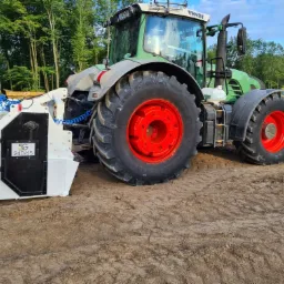 Ciągnik Fendt z frezarką do pni na terenie budowy, w tle drzewa i wywrotka.