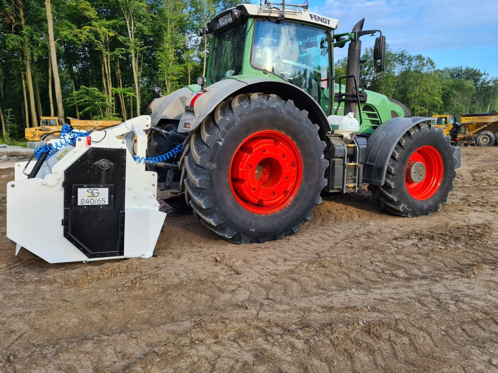Ciągnik Fendt z frezarką do pni na terenie budowy, w tle drzewa i wywrotka.