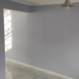 Świeżo pomalowane na szaro ściany w pomieszczeniu, widoczna lampa sufitowa i fragment podłogi z jasnych płytek, wpadające przez okno światło.