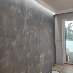 Ściana z efektem dekoracyjnym w odcieniach szarości, oświetlenie LED podsufitowe, widoczne puszki elektryczne przed montażem osprzętu, fragment podłogi z paneli oraz okno balkonowe.