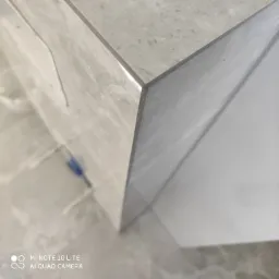 Detal narożnika obudowy umywalki z szarych, matowych płytek ceramicznych, z widocznymi fugami i fragmentem podłogi wyłożonej podobnymi płytkami.