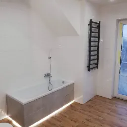 Nowoczesna łazienka z białą wanną obudowaną szarą płytą, podświetleniem LED u dołu, białymi płytkami na ścianach z delikatnym wzorem fal, drewnopodobną podłogą i czarnym grzejnikiem drabinkowym.