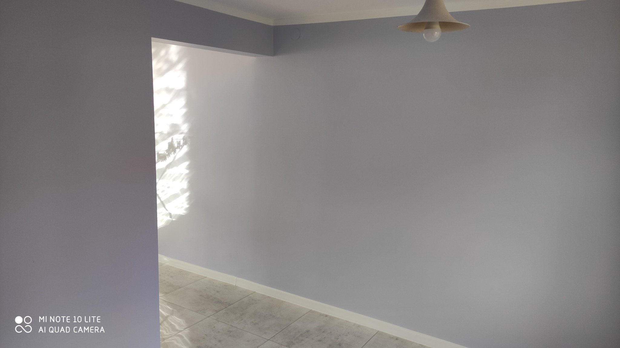 Świeżo pomalowane na szaro ściany w pomieszczeniu, widoczna lampa sufitowa i fragment podłogi z jasnych płytek, wpadające przez okno światło.