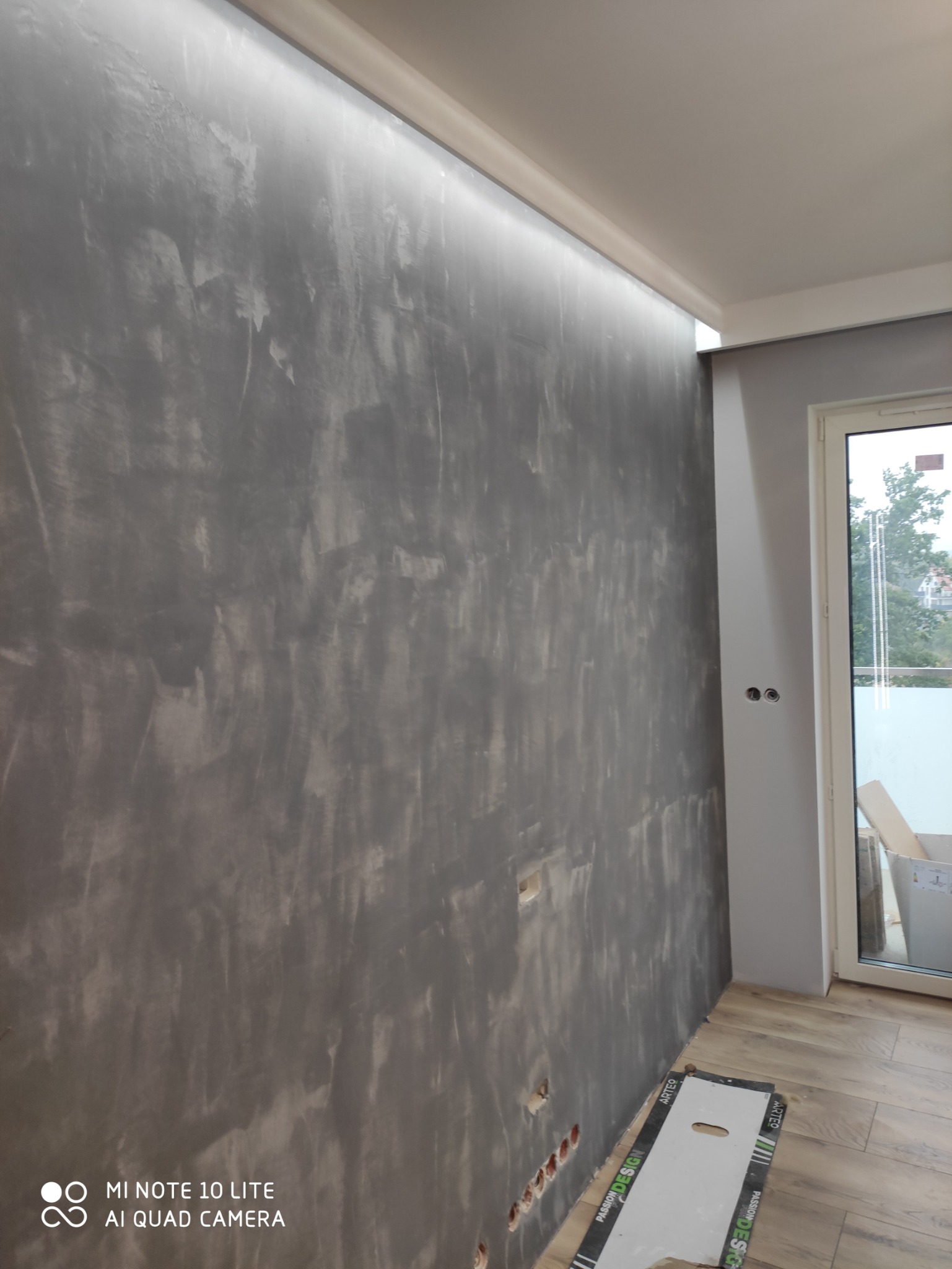 Ściana z efektem dekoracyjnym w odcieniach szarości, oświetlenie LED podsufitowe, widoczne puszki elektryczne przed montażem osprzętu, fragment podłogi z paneli oraz okno balkonowe.